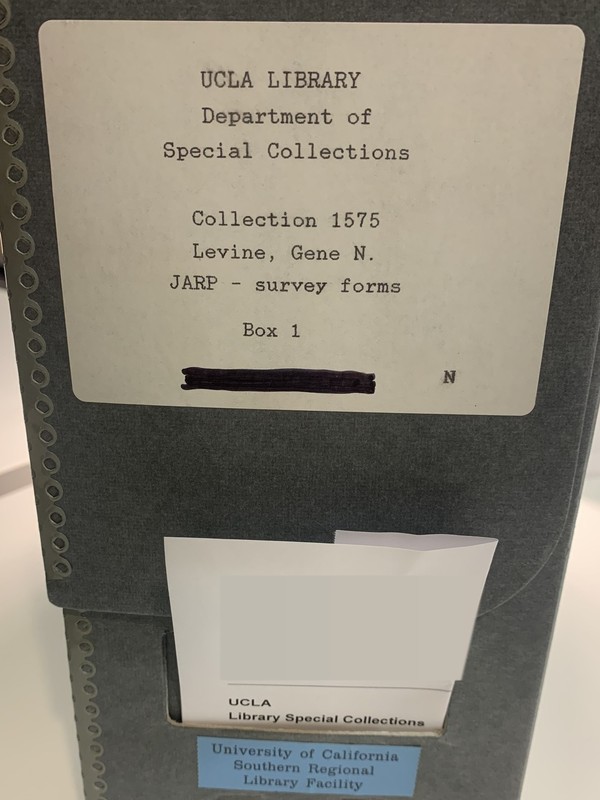 A gray archive box standing on a table, labeled UCLA Library, Department of Special Collections, Collection 1575, Levine, Gene N., JARP - survey forms, Box 1.